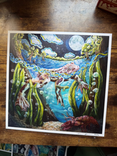 Load image into Gallery viewer, A square art print featuring a colorful fantasy illustration of a river flowing into the sea, with sea creatures like a lobster and fish, set on a wooden tabletop.
